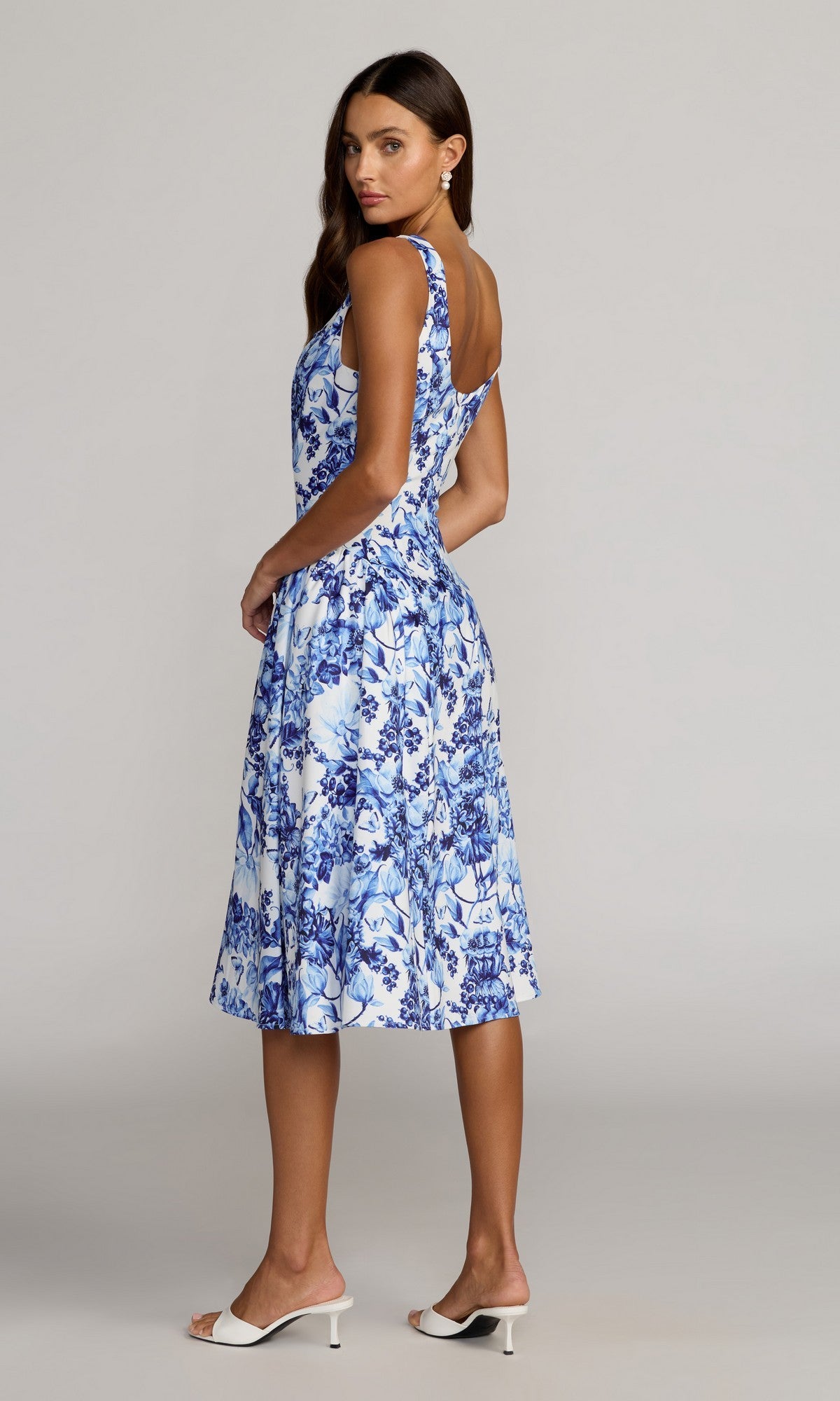 A woman stands sideways against a plain background wearing the Mac Duggal 37155 Floral Midi Wedding Guest Dress, which features a sleeveless square neckline in white and blue floral print, paired with white heeled sandals.